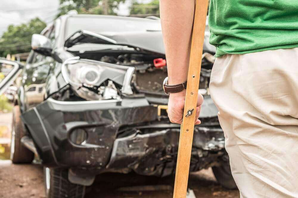 Injured driver in crutches in standing in front of damage car from collision.